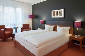 A hotel room with a large bed, two bedside tables, a chair, a small table with a lamp, and a window with sheer curtains. - In the heart of Erfurt - double room at Hotel Dorint (Erfurt)