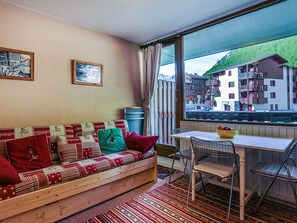 Um quarto aconchegante com um sofá de madeira, uma mesa de jantar e cadeiras. Há duas fotos emolduradas na parede e uma janela com vista para edifícios.