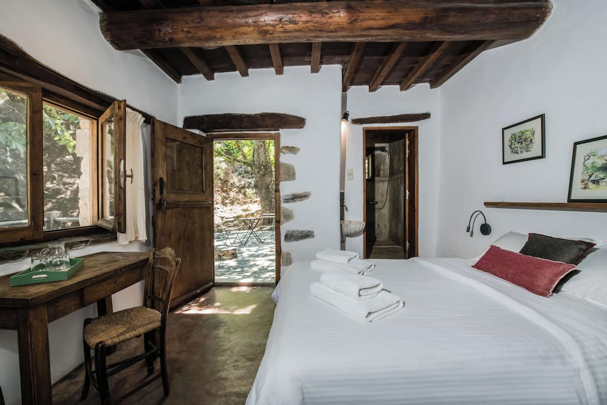 A bedroom with a wooden desk, a chair, a bed with white linens, and a stone wall.