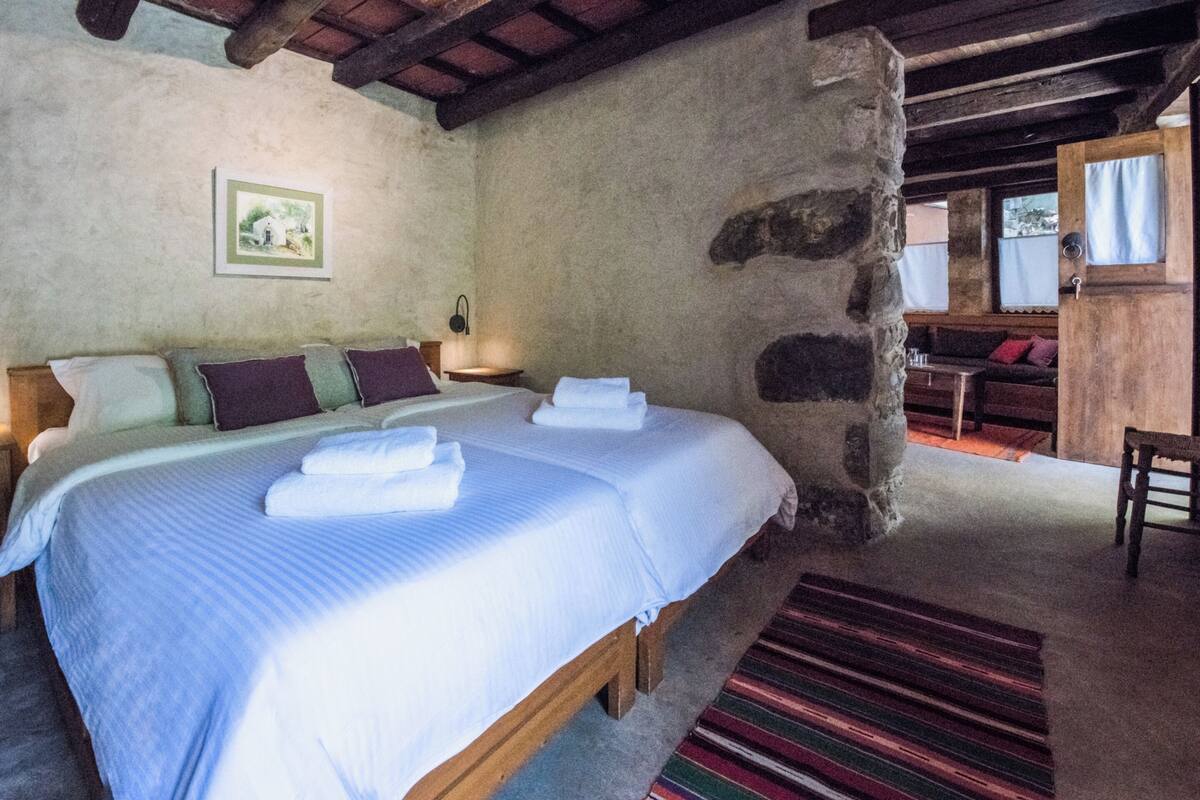 A bedroom with a large bed, a stone wall, a wooden bench, and a view of the outside.