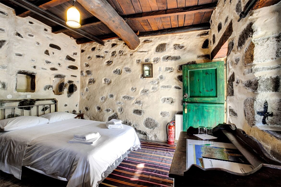 A rustic room with a stone wall, a wooden ceiling, a bed with white linens, a green cabinet, and a wooden bench.