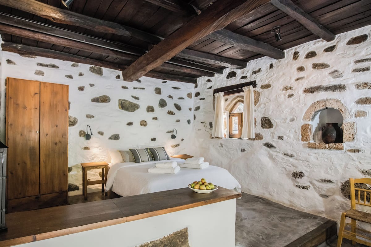 A rustic bedroom with a stone wall, wooden beams, a bed with white linens, a wooden wardrobe, and a small table with fruit.