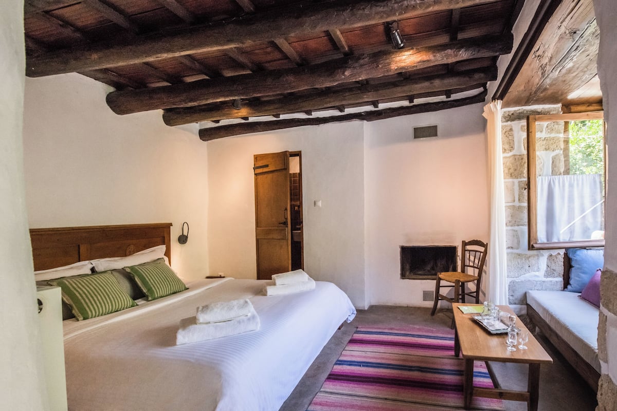 A bedroom with a wooden ceiling, a bed with green striped pillows, a wooden table with glasses, and a stone wall.