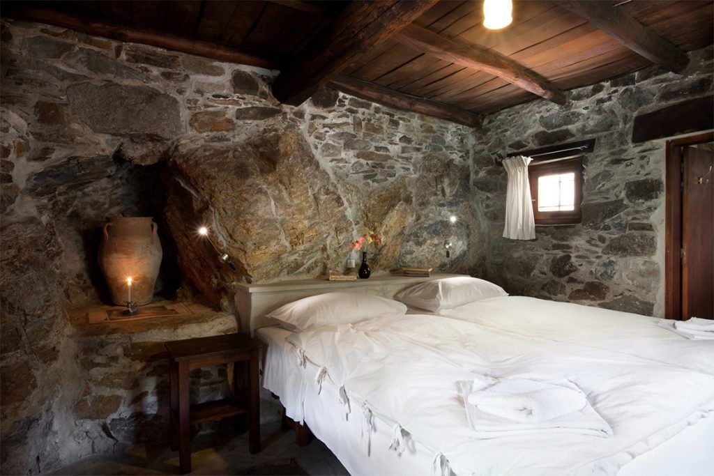 Une pièce rustique aux murs de pierre, avec un lit, une petite table sur laquelle est posée une bougie, et une fenêtre ornée de rideaux.