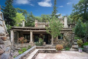 Un bâtiment rustique en pierre, doté d’un porche en pierre, entouré d’une végétation luxuriante et d’arbres.