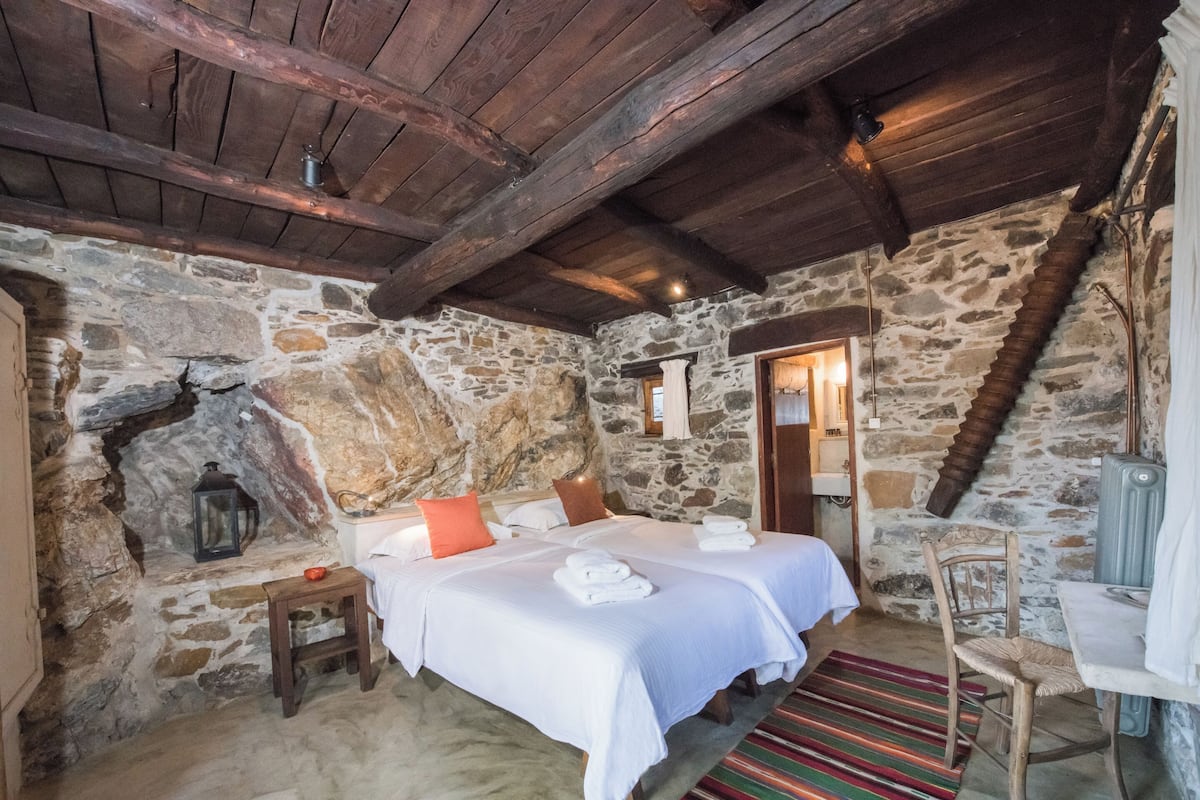 A rustic stone-walled bedroom with two beds, wooden beams, a stone fireplace, and a small table with a lantern.