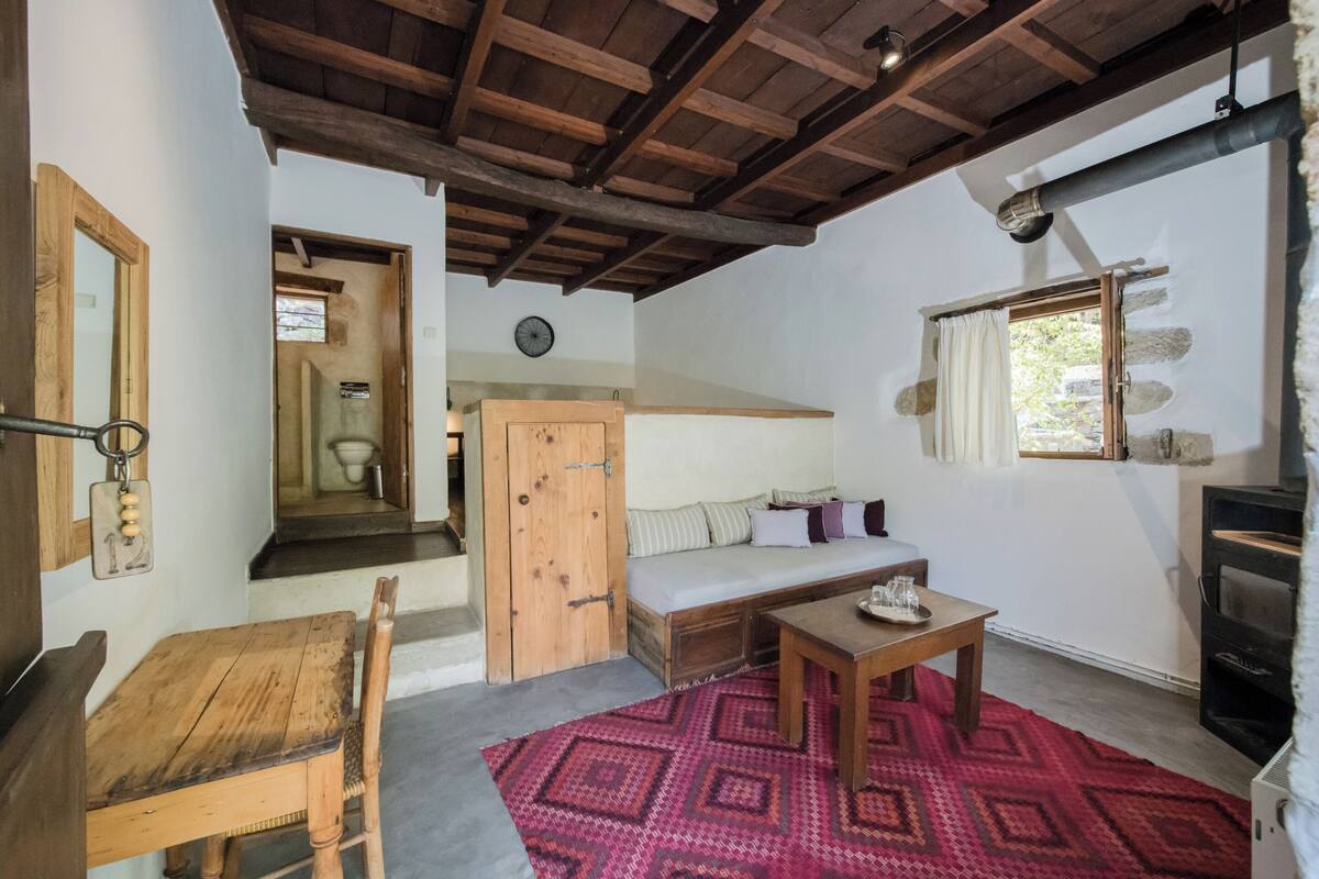A rustic room with a wooden table, a sofa, a red patterned rug, a fireplace, and a wooden ceiling.