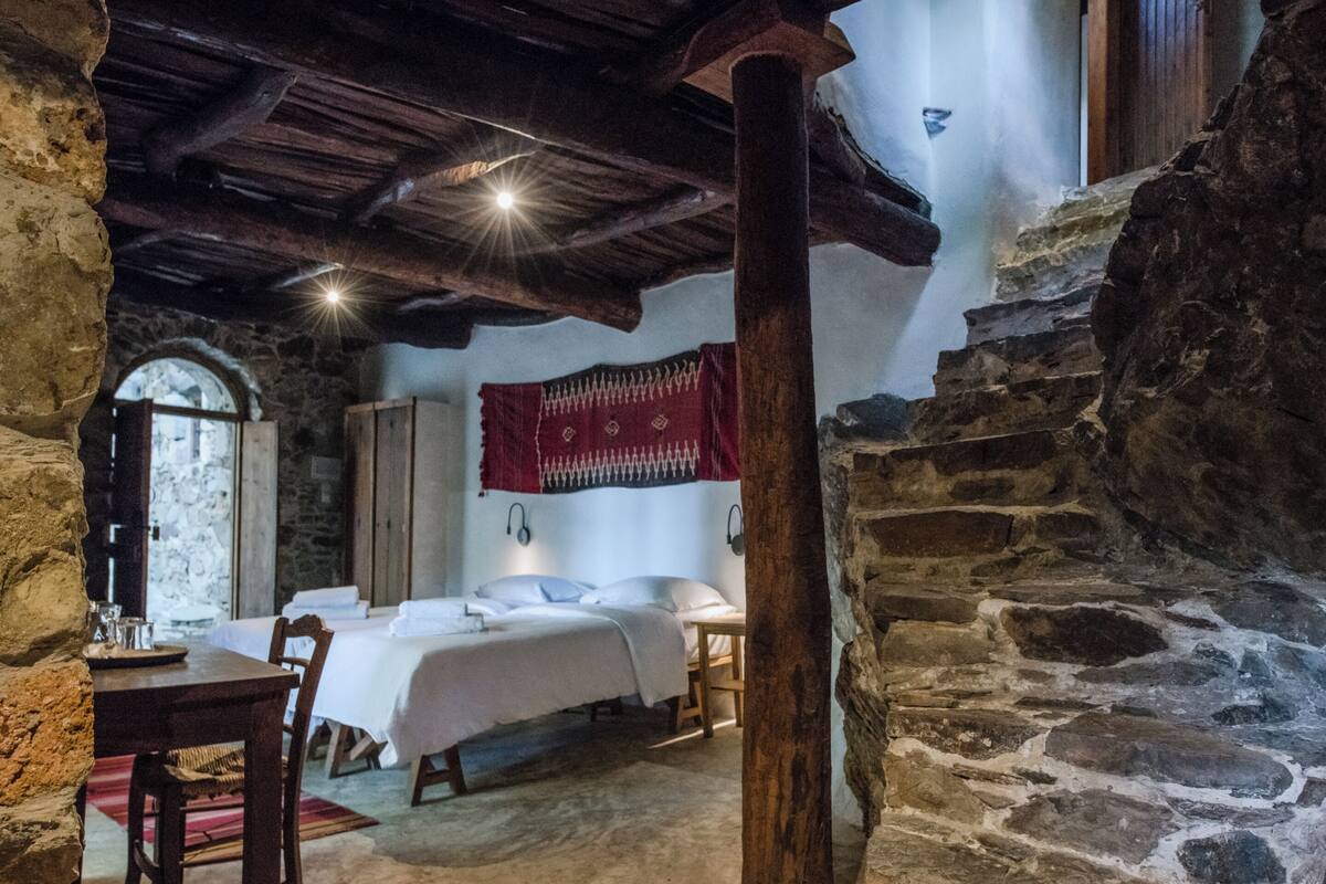 A rustic room with stone walls, wooden beams, a bed, a table, and a chair.