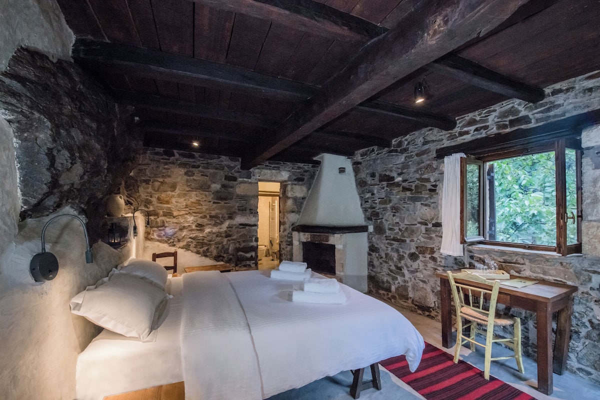 A rustic room with a stone wall, a bed with white linens, a wooden table, and a fireplace.
