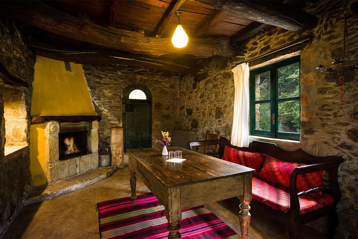 A rustic stone room with a wooden table, a fireplace, a red sofa, and a window with a view of greenery.