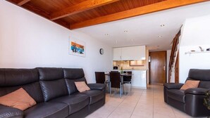 Ein modernes Wohnzimmer mit zwei schwarzen Ledersofas, einem Esstisch mit Stühlen, einer Küche im Hintergrund und einer Holzbalkendecke.