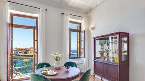 Ein Essbereich mit einem runden Tisch, grünen Stühlen, einer Vitrine mit Glastüren und Meerblick durch große Fenster.