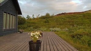 A wooden deck with a potted plant, a building with large windows, and a grassy landscape with a hill in the background. - Odinsve Country Home (Selfoss)