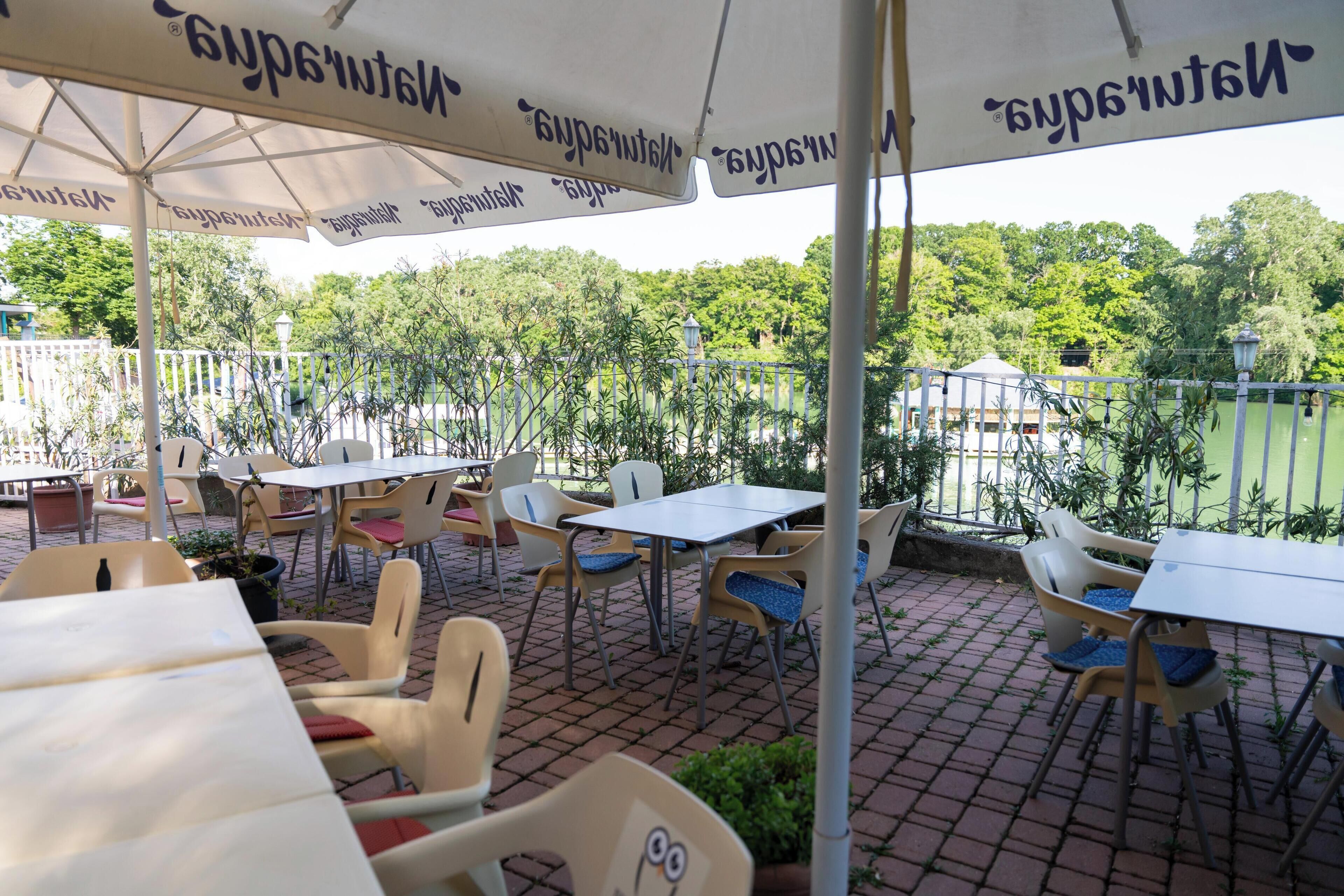 An outdoor patio area with tables and chairs, umbrellas, and a view of a green park.