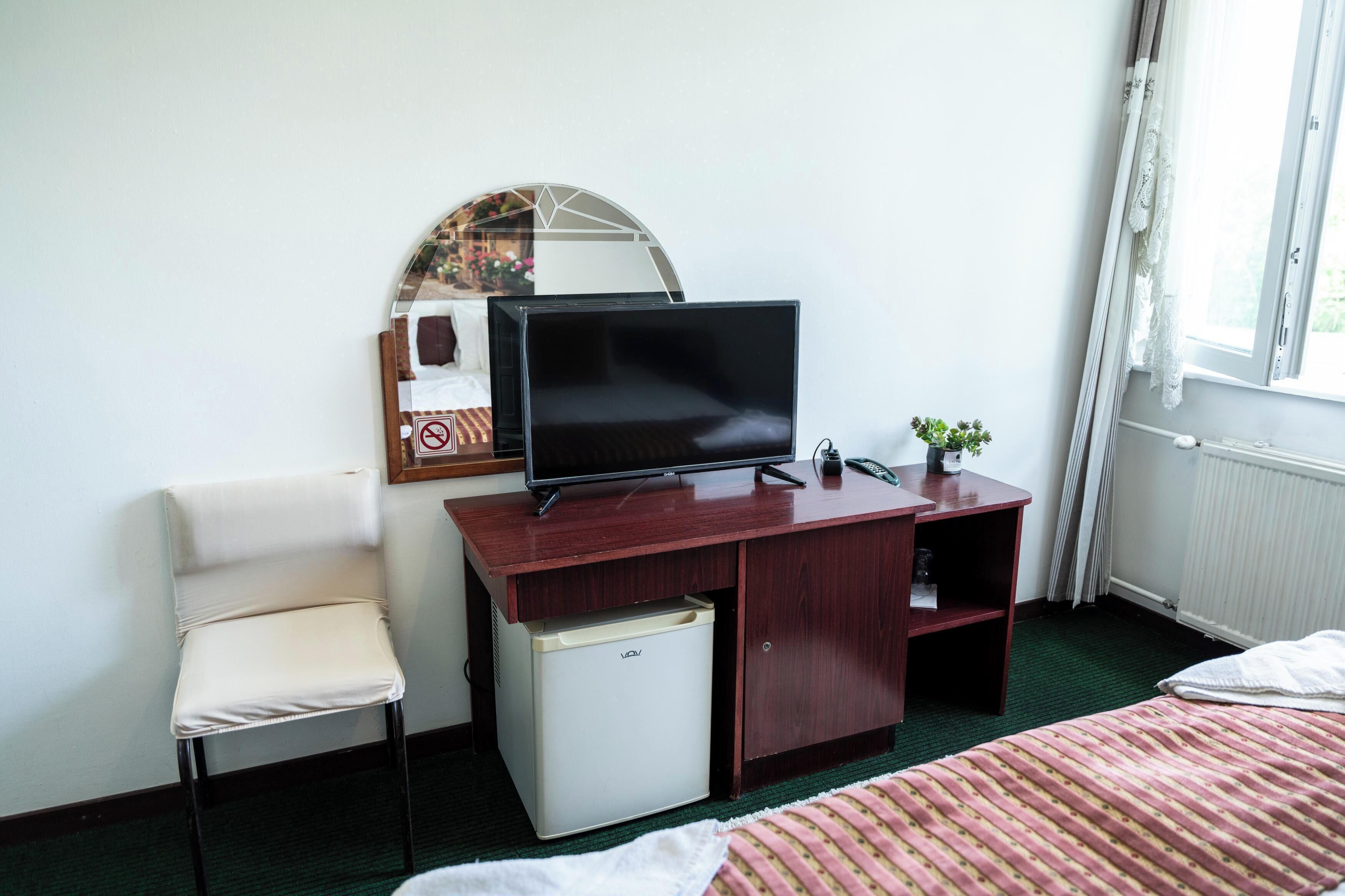 A hotel room with a bed, a desk with a television, a chair, a small refrigerator, and a window with curtains.