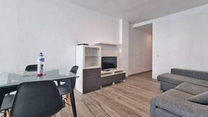 A modern living room with a glass dining table, black chairs, a grey sofa, a TV on a white cabinet, and a water bottle on the table. - Costa Azul - 3 Left (Puerto de Naos)