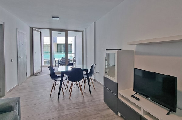 Una sala de estar moderna con una televisión de pantalla plana, una mesa de comedor con sillas y un ventanal que da a un espacio al aire libre.
