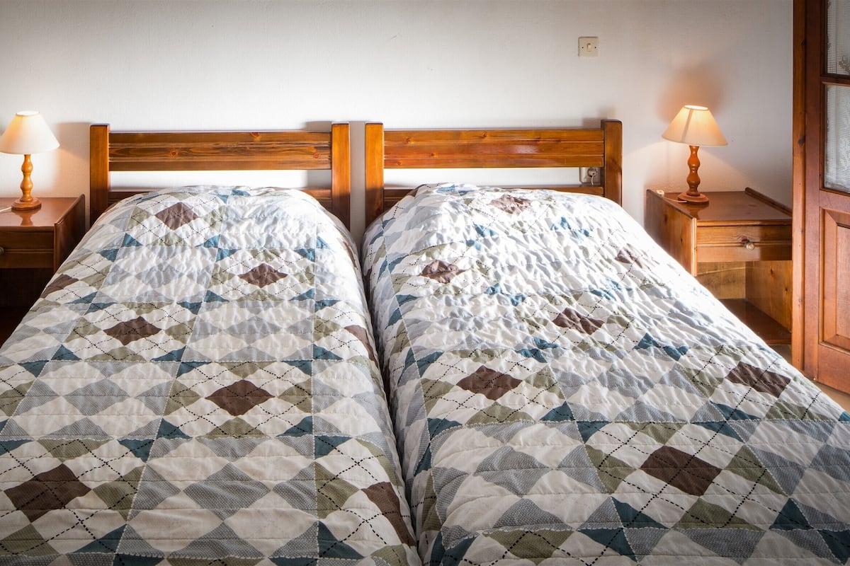A bedroom with two single beds, wooden headboards, bedside tables with lamps, and a wooden wardrobe.