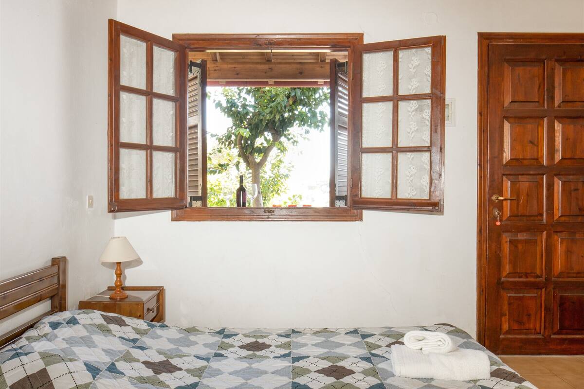 A bedroom with a wooden bed, a nightstand with a lamp, a window with a view of trees, and a wooden door.