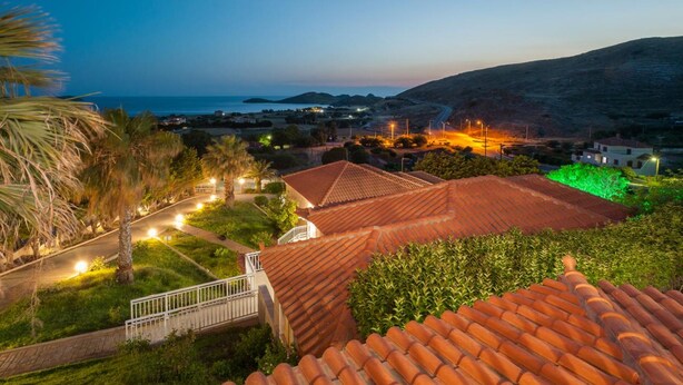 A coastal town at dusk with terracotta-roofed buildings, a paved road, and a view of the sea. - Iliovasilema (Lemnos)