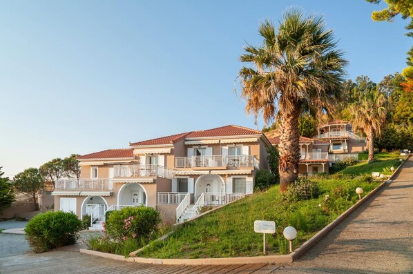 A two-story building with a red-tiled roof, multiple balconies, and a landscaped garden with palm trees.