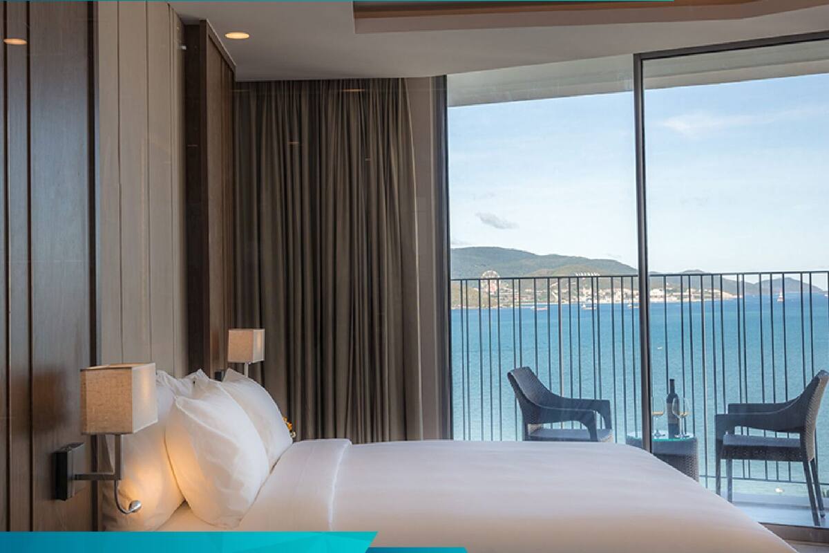 A hotel room with a large bed, a view of the sea, and a balcony with chairs.