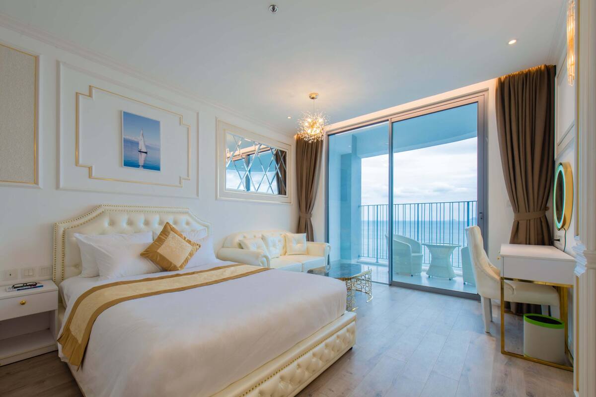 A spacious bedroom with a large bed, a sitting area, and a balcony with a view of the sea.