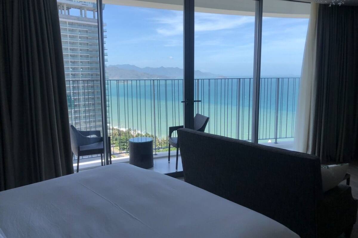 A hotel room with a large bed, a small table, two chairs, and a view of the ocean through floor-to-ceiling windows.