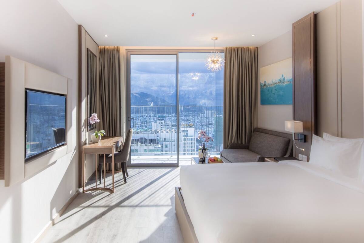 A modern hotel room with a large bed, a desk, a chair, and a view of the city through floor-to-ceiling windows.