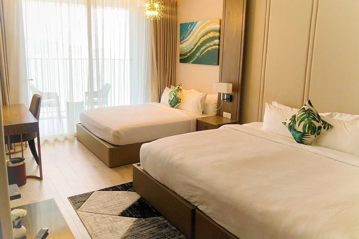 A hotel room with two beds, a desk, a chair, and a large window with sheer curtains.