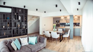 Een moderne woonkamer met een grijze bank, witte stoelen, een eettafel en een keukenhoek met een trap.