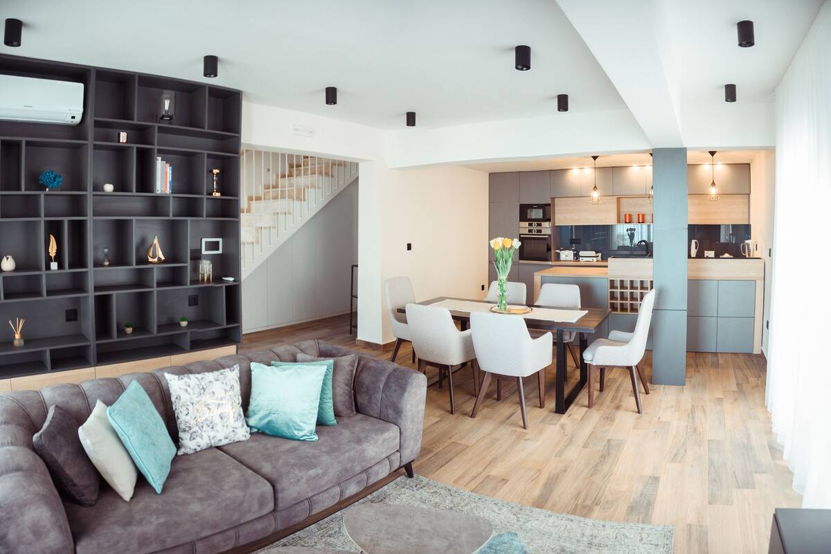 Ein modernes Wohnzimmer mit grauem Sofa, weißen Sesseln, einem Esstisch und einer Küchenzeile mit Treppe.