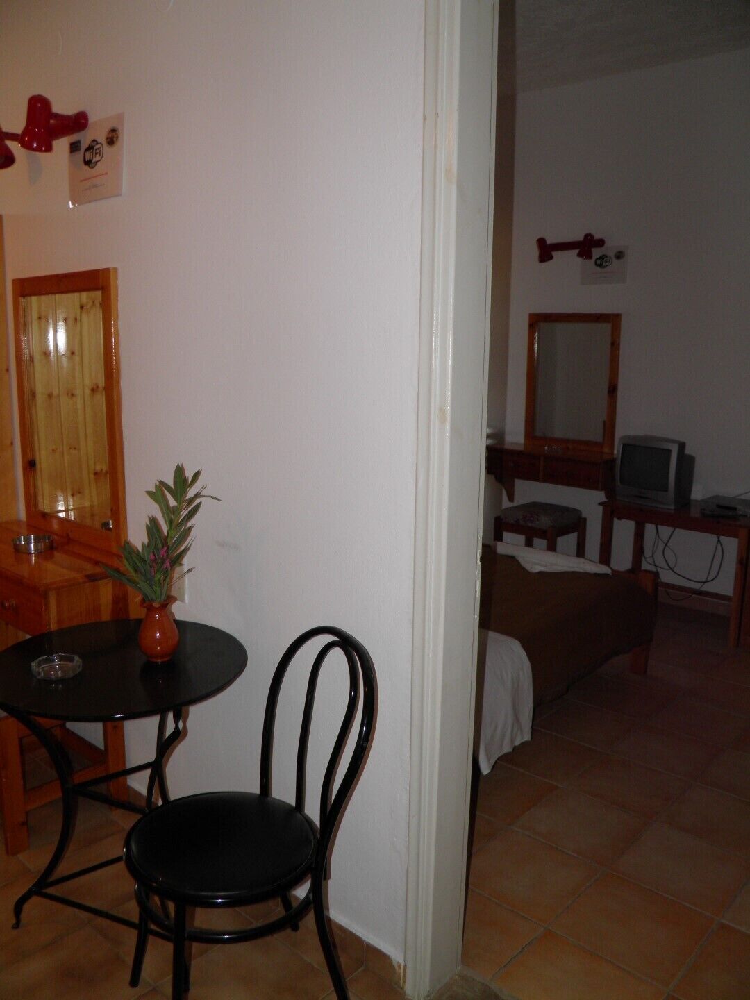 Una habitación con puerta de madera, una mesita con un jarrón, una silla negra, una cama, un televisor y un espejo en la pared.