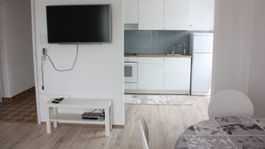Una habitación moderna con un televisor de pantalla plana en la pared, una pequeña mesa blanca con un control remoto, y vistas a una cocina con armarios blancos y un refrigerador.