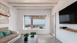 A modern living room with a large window, a flat-screen TV, a sofa, and a coffee table with books and a plant. - Infinity View and SUNSET Over Aegean Sea, PRIVCY & Security (Mykonos)
