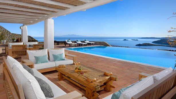 A poolside area with a wooden table, white sofas, and lounge chairs overlooking a clear blue sea and distant mountains. - Infinity View and SUNSET Over Aegean Sea, PRIVCY & Security (Mykonos)