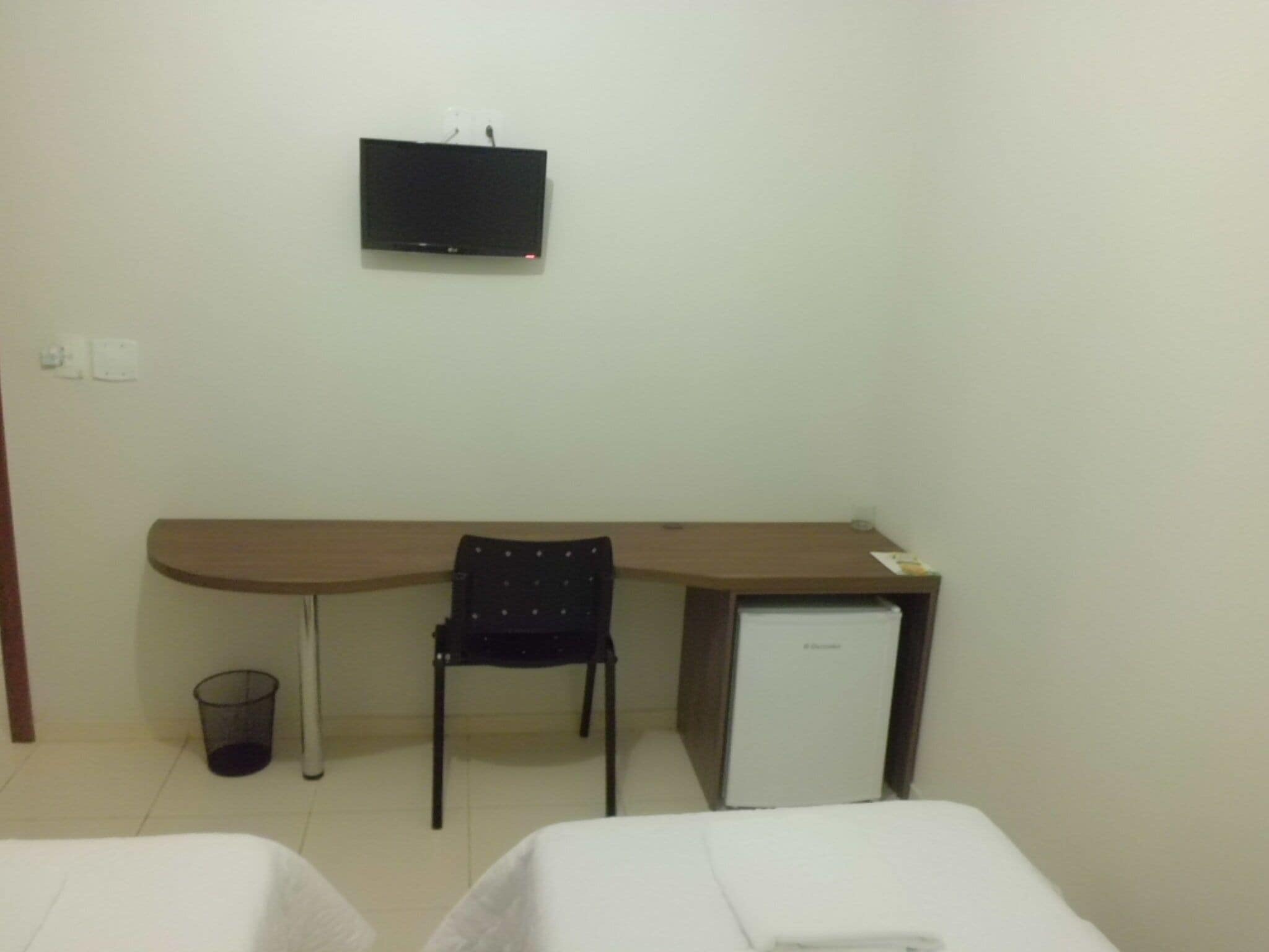 A hotel room with a desk, a chair, a small refrigerator, and a TV mounted on the wall.
