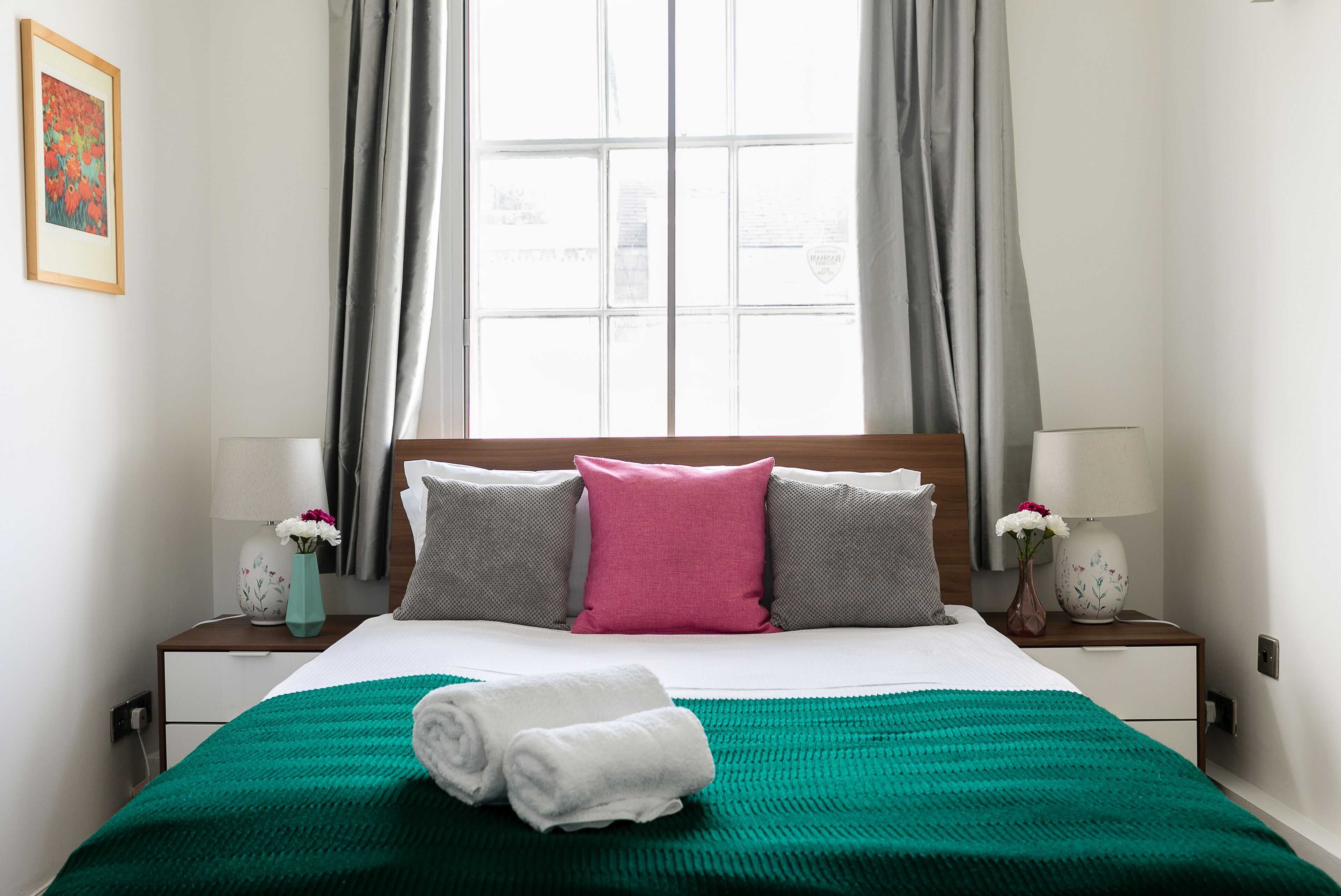 A neatly made bed with a green blanket, two pillows, and a rolled towel. There are two bedside tables with lamps and vases of flowers.