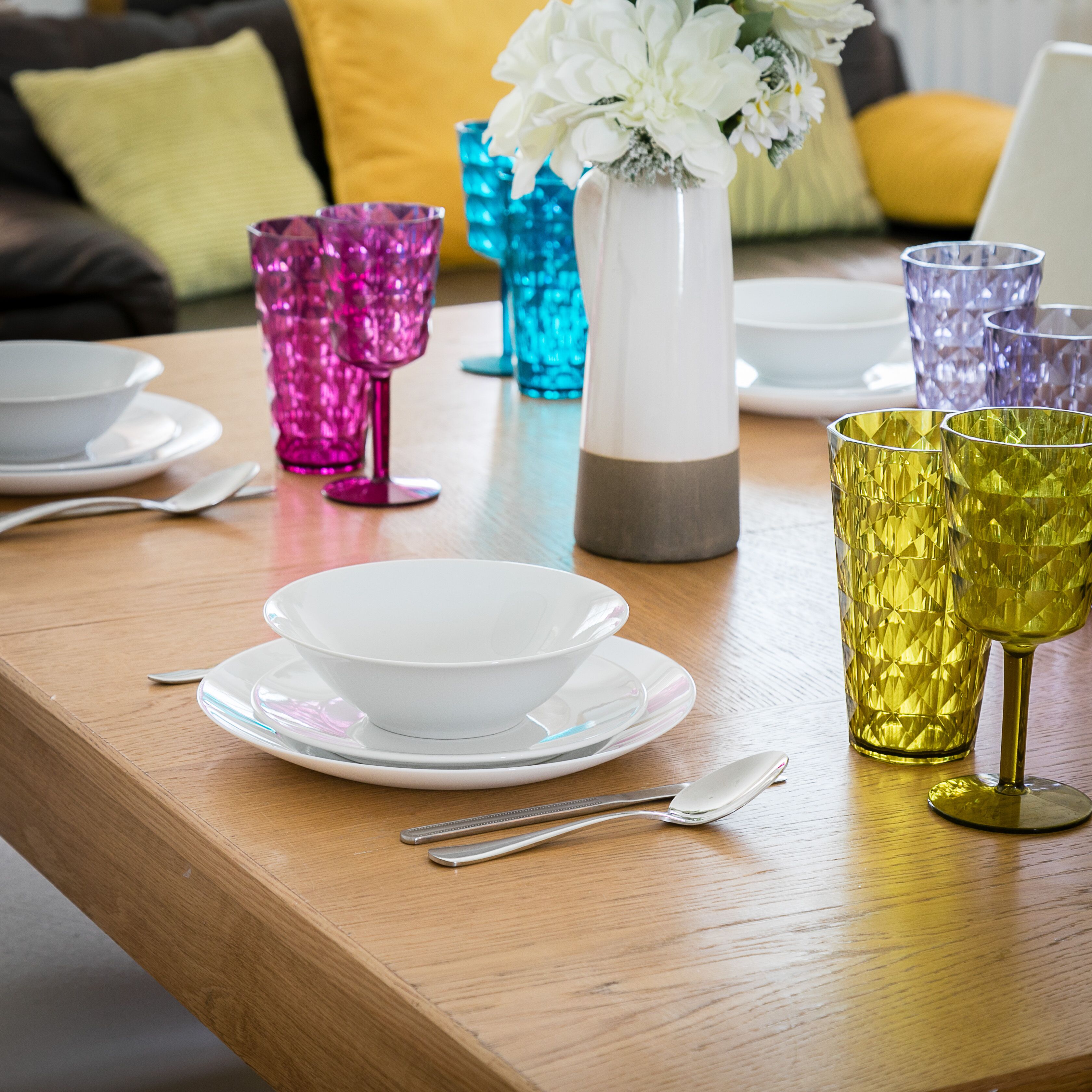 A dining table with a white bowl, two spoons, and colorful glassware.