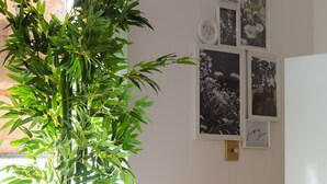 Ein Esstisch ist gedeckt mit Tellern, Schüsseln, Gläsern und Besteck. Eine große, grüne Pflanze steht in einer Vase, und an der Wand hängen gerahmte Bilder.