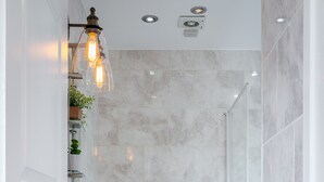 Ein modernes Badezimmer mit einem weißen Waschbecken, einer Toilette und einer Dusche. Es gibt Regale mit Pflanzen und eine Leuchte über dem Waschbecken.