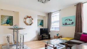 Een moderne woonkamer met een glazen salontafel, een bank en een televisie. Er hangen schilderijen aan de muur en een raam met gordijnen.