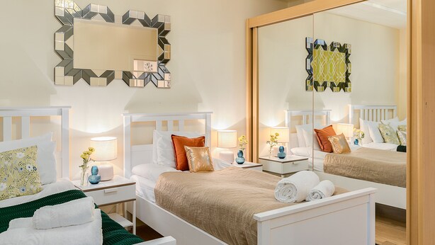 Een tweepersoonsbed met een beige sprei, twee nachtkastjes met lampen, een grote spiegel en een decoratief wandobject.