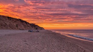 A beach with a sunset, sandy shore, and a few people in the distance. - Beautiful Spacious Family Retreat w/ Pool! (Eastham)