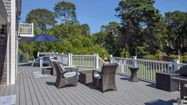 A wooden deck with outdoor furniture, including wicker chairs and a table, surrounded by white railings and trees. - Beautiful Spacious Family Retreat w/ Pool! (Eastham)