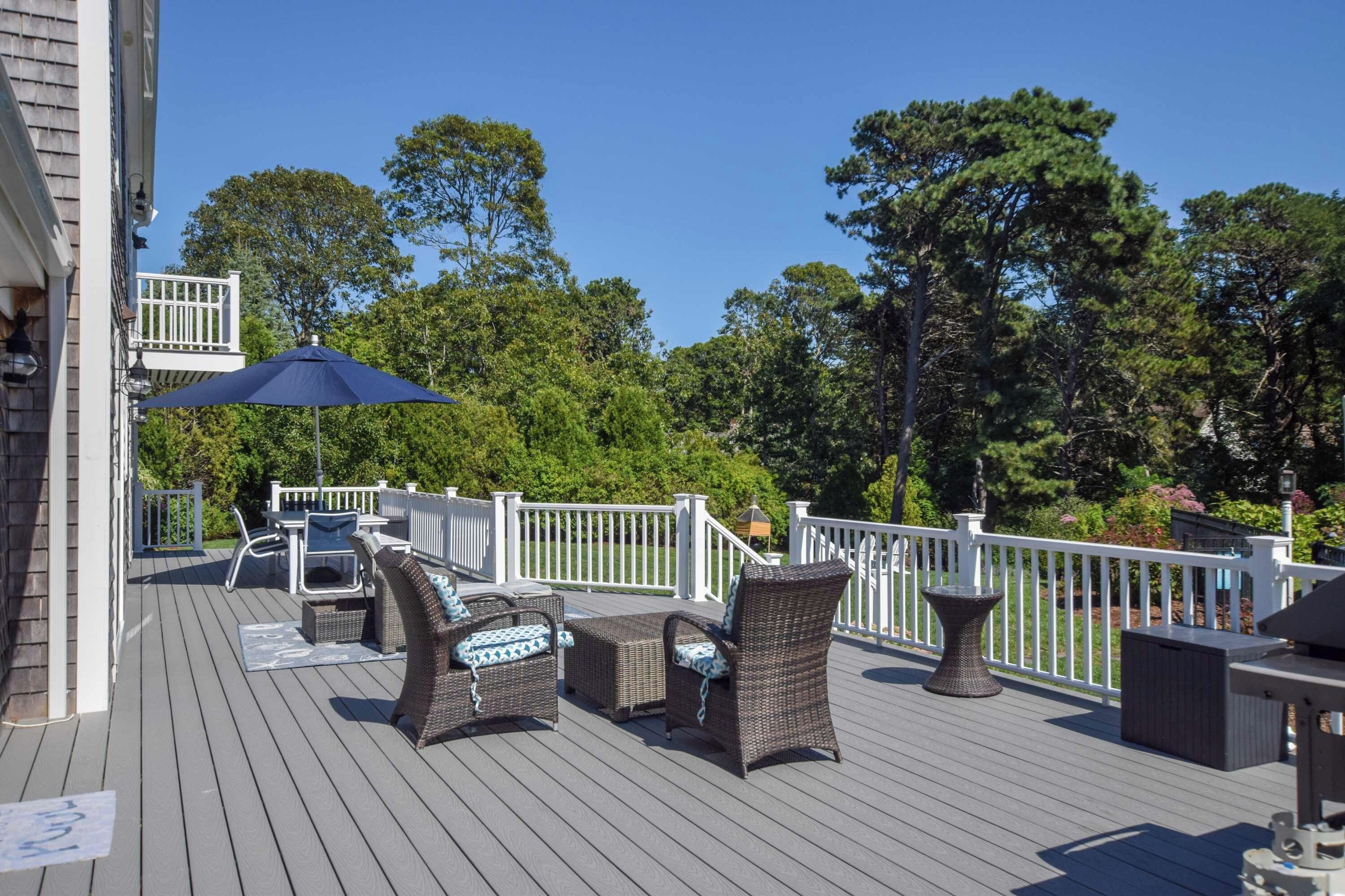 Eine Holzterrasse mit Gartenmöbeln, darunter Korbstühle und ein Tisch, umgeben von weißen Geländern und Bäumen.