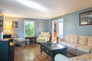 A living room with a sofa, armchair, ottoman, and a coffee table. There is a window with a view of greenery outside.