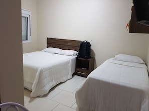 A room with two single beds, a wooden nightstand with a black bag, and a window with blinds.