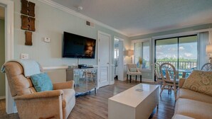 A living room with a sofa, a chair, a coffee table, a TV, and a view through the window. - Family Paradise- 2bdrm/2 bath condo, indoor pool, spa, outdoor pools, & tennis. (Hilton Head Island)