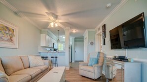 A living room with a sofa, armchair, and coffee table, a kitchen area with cabinets and a TV, and a ceiling fan. - Family Paradise- 2bdrm/2 bath condo, indoor pool, spa, outdoor pools, & tennis. (Hilton Head Island)